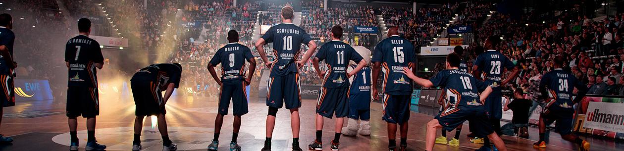 Des joueurs de basket-ball en maillots bleus avec des numéros et des noms au dos se tiennent sur le terrain devant un public.