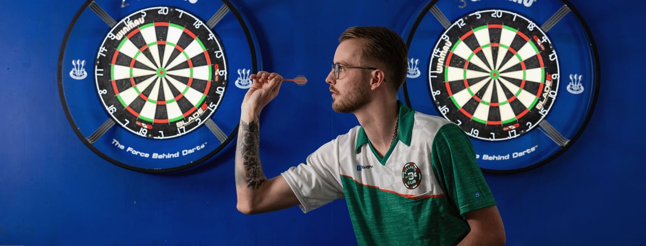 A man with glasses is shown in profile, aiming a dart at a dartboard that is not visible in the image. The background consists of a blue wall with two dartboards hanging on it.