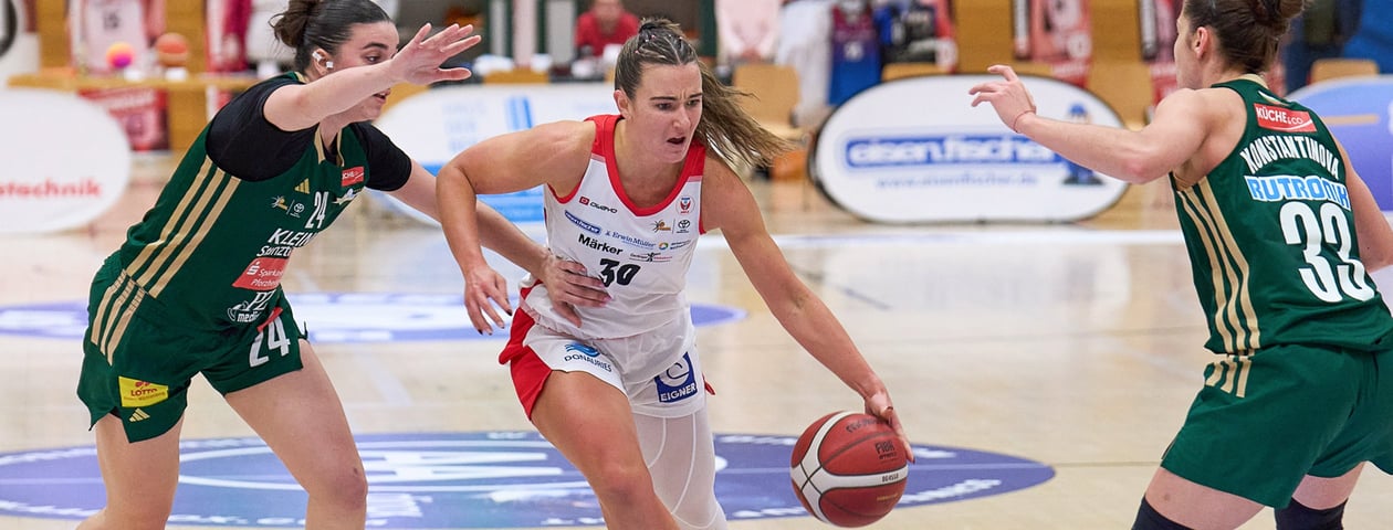 Three women play basketball, one in a white jersey with red accents dribbles the ball while two opponents in green jerseys defend.