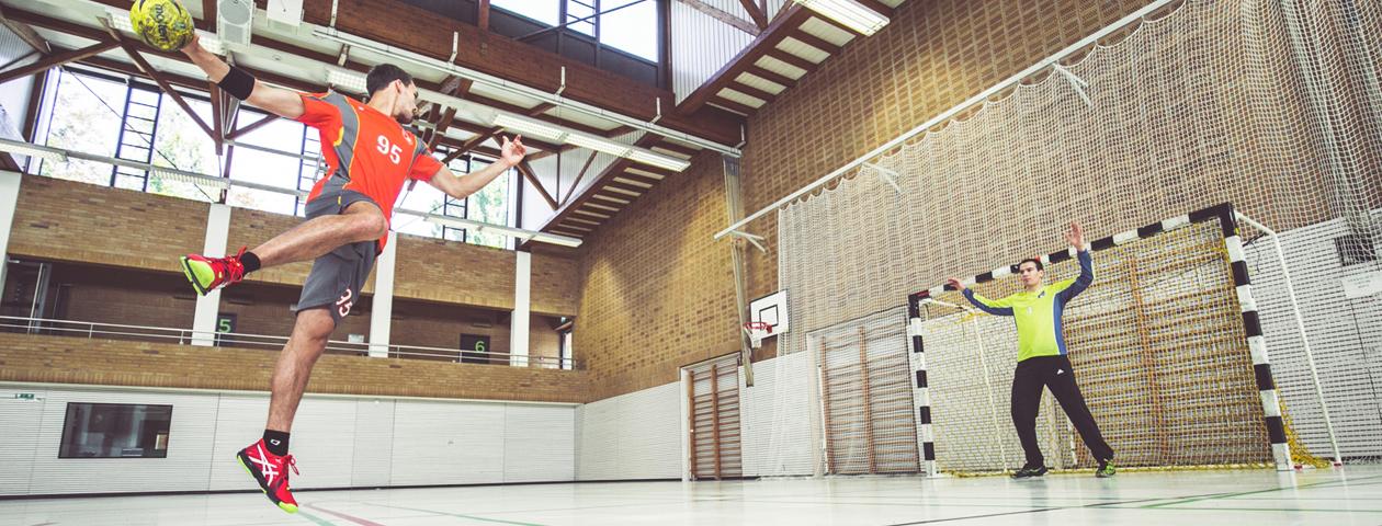 A man is playing handball in a hall while a woman stands in the background with her arms raised. The clothing is from the brand 'owayo'.