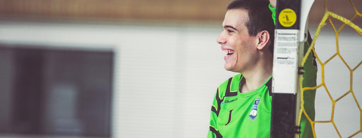 A man wearing a green handball jersey with customization options from owayo smiles while standing in front of a goal net.