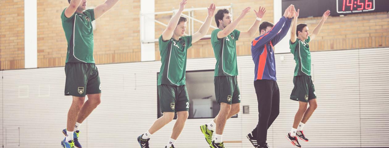 A group of people wearing green handball jerseys is jumping indoors. A coach in an orange training suit stands in front of them.