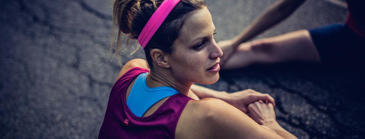 Een vrouw draagt een kleurrijk hardloopshirt met een roze hoofdband en zit op de grond tijdens een pauze bij het joggen.
