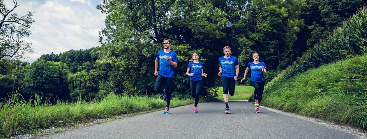 Vier mensen lopen op een landweg, allemaal met blauwe hardloopshirts die het woord 'owayo' dragen. De achtergrond toont een groene bos en een heldere zomerdag.