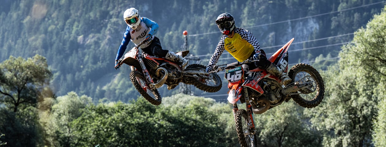 Zwei Motorradfahrer fliegen durch die Luft, während sie auf Motocross-Höhenflug-Schutzanzügen in der Luft springen. Der Hintergrund zeigt eine natürliche Landschaft mit Bäumen und einem bewaldeten Hügel.