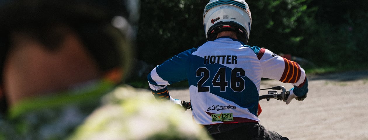 Ein Motocross-Fahrer trägt ein selbstgestaltetes Trikot mit der Nummer 246 und dem Namen 'HOTTER' auf dem Rücken. Das Bild zeigt den Fahrer von hinten während er eine Strecke fährt.
