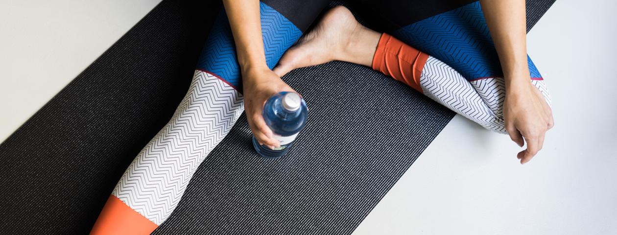 Una persona está sentada en una estera de yoga y sostiene una botella de agua en sus manos. Lleva pantalones de yoga coloridos y personalizados.