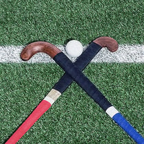 Dos palos de hockey cruzados diagonalmente en un campo de juego. En medio, en un lado, hay una pelota de hockey sobre césped.