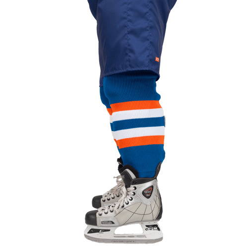 Seitenansicht eines Eishockeyspielers mit blauen Hosen, gestreiften Socken in Blau, Orange und Weiß sowie Eishockeyschuhen.