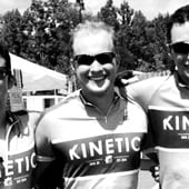 A man is wearing a cycling jersey with the text 'KINETIC' and standing in front of a group of people. The background shows trees.