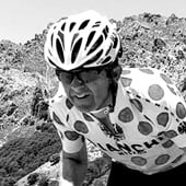 A cyclist wearing a shirt with large white dots on a black background is riding through a hilly landscape.