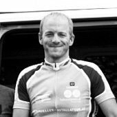A man wearing a cycling jersey with the logo of 'owayo' stands in front of a vehicle. The image is black and white.