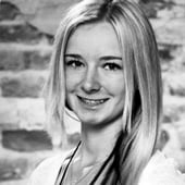 A woman with blonde hair stands in front of a wall wearing a shirt.