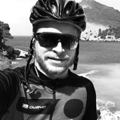 A man wearing a bicycle helmet and an owayo jacket stands in front of a coastal landscape.