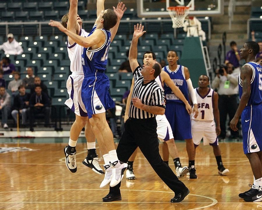 Ein Basketballspiel in einer Halle mit einem Schiedsrichter in der Mitte, der die Spieler beobachtet, während zwei Spieler in blauen und weißen Trikots springen, um den Ball zu erreichen.
