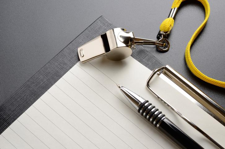 Ein silberner Pfiff mit gelber Schnur, ein Kugelschreiber und ein Notizblock auf einem grauen Untergrund.
