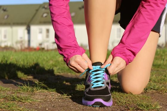 Eine Person in einer pinken Laufjacke kniet auf dem Rasen und bindet die Schnürsenkel eines schwarzen und lila Laufschuhs mit blauen Schnürsenkeln.