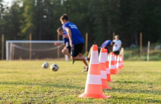 Zwei junge Männer in blauen Trikots sprinten auf einem Fußballfeld, während sie um orange-weiße Hütchen dribbeln.