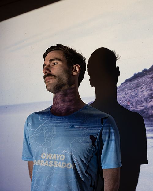 Een man draagt een blauw gestreept T-shirt met de tekst 'Owayo Ambassador' en staat voor een achtergrond die een landschap toont.
