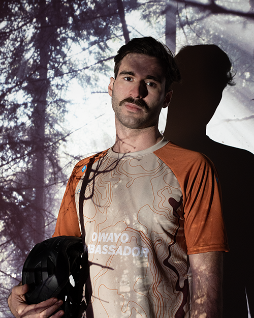 Een man met een t-shirt waarop 'owayo ambassador' staat, staat in het bos en houdt een helm vast.