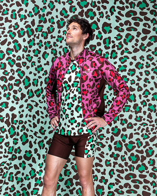 Een man draagt een roze shirt met luipaardprint en bruine broek tegenover een achtergrond die eveneens een luipaardprint heeft.