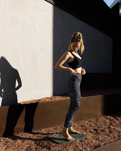 Une femme porte un haut noir et des pantalons à rayures, debout sur une tapis de yoga. Son ombre est visible sur le mur.