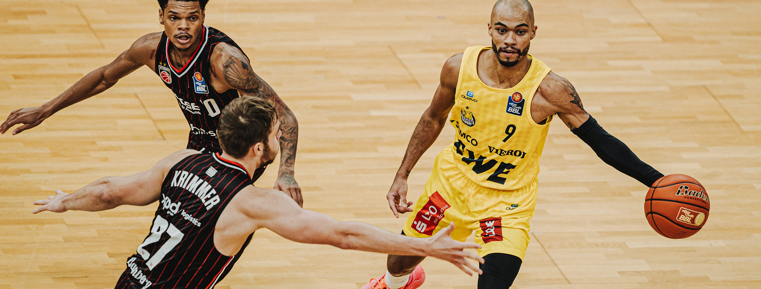 Ein Basketballspieler in einem roten Owayo-Trikot während eines Spiels, mit einem Basketball in der Hand und einem Gegner in der Verteidigung.