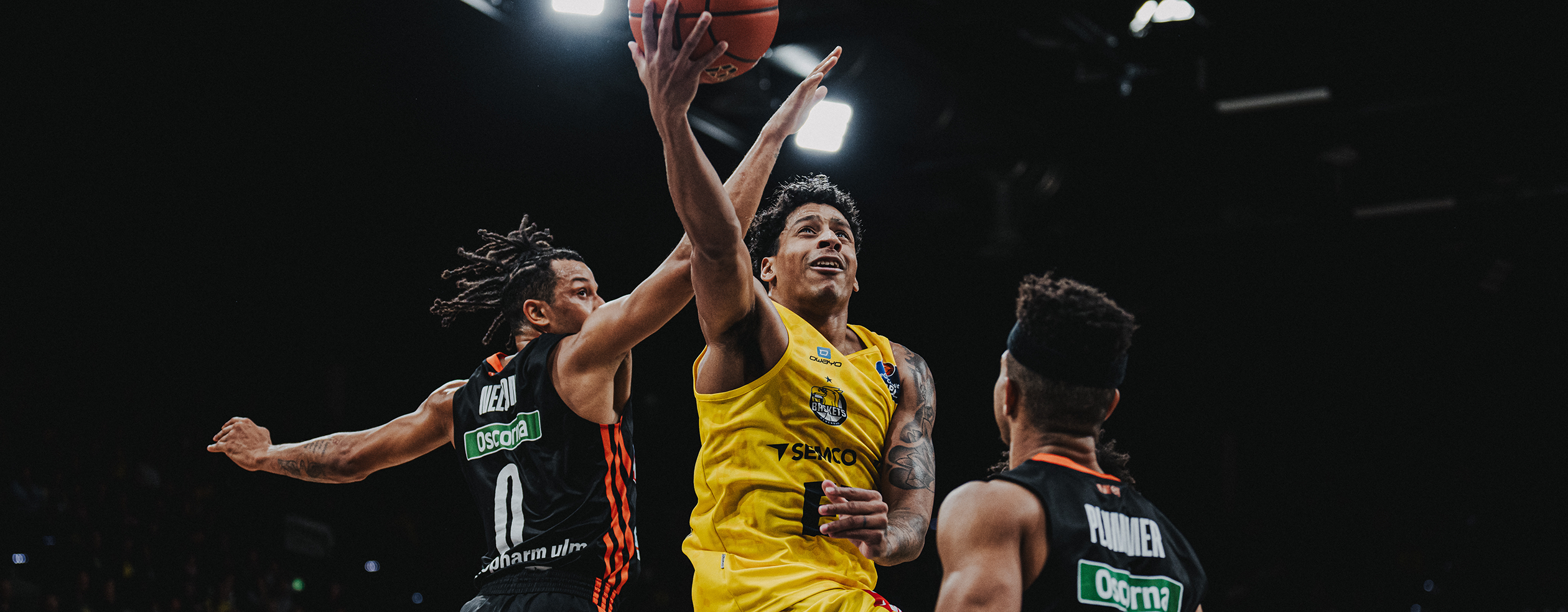 A basketball player in a yellow Owayo jersey dribbling while an opponent in a black jersey tries to defend him