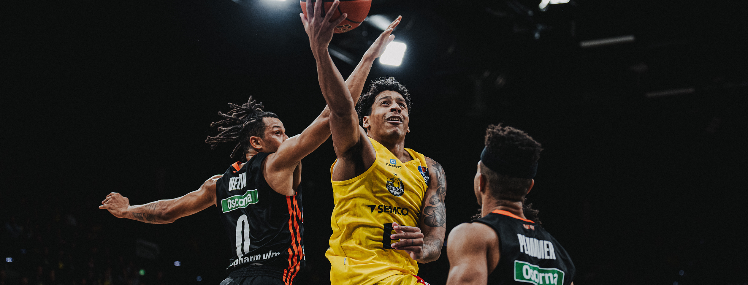 A basketball player in a yellow Owayo jersey dribbling while an opponent in a black jersey tries to defend him