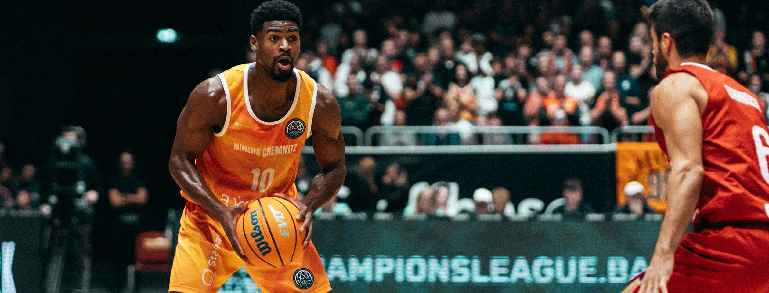 Zwei Basketballspieler in orangefarbenen Owayo-Trikots während eines Spiels. Einer dribbelt den Basketball, während der andere mitläuft.