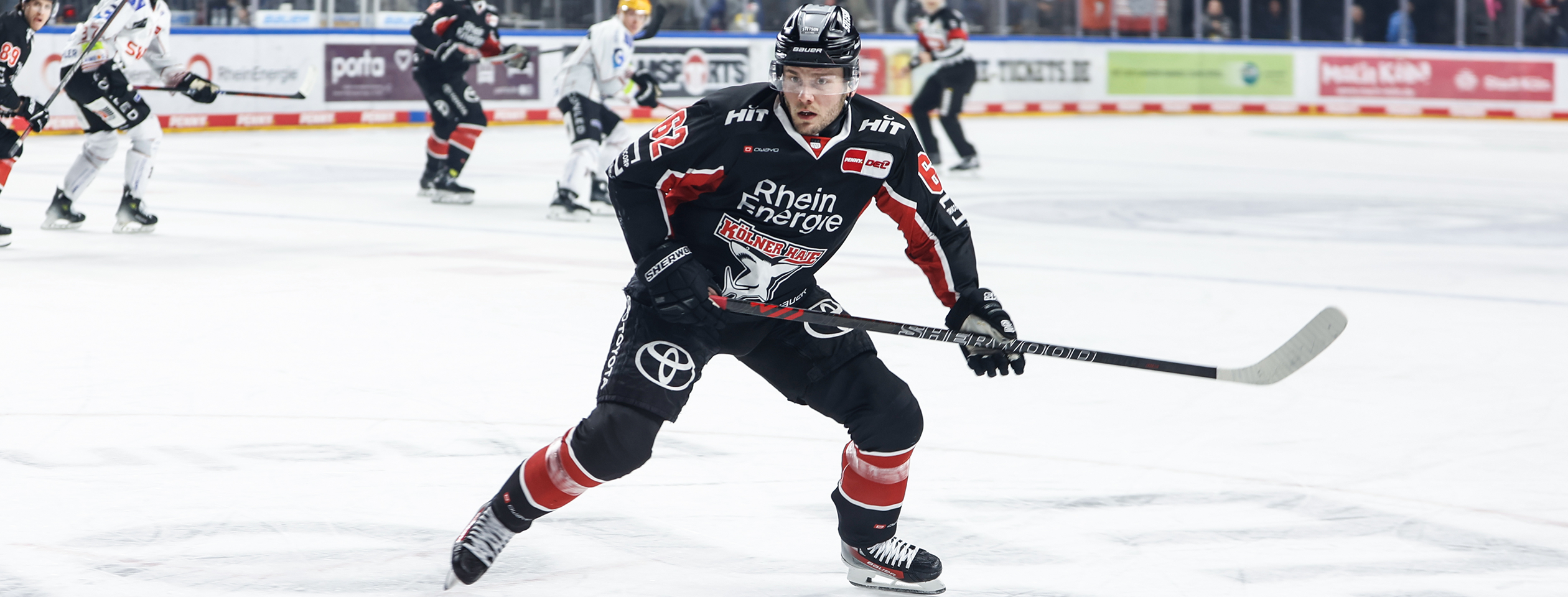 An hockey player in a red Owayo jersey controls the puck on the ice, while an opponent in a white jersey follows him.