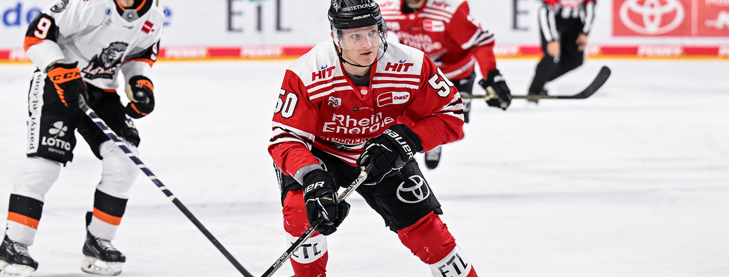 An hockey player in a black Owayo jersey skates across the ice with his stick, while other players in white and black jerseys are in the background.