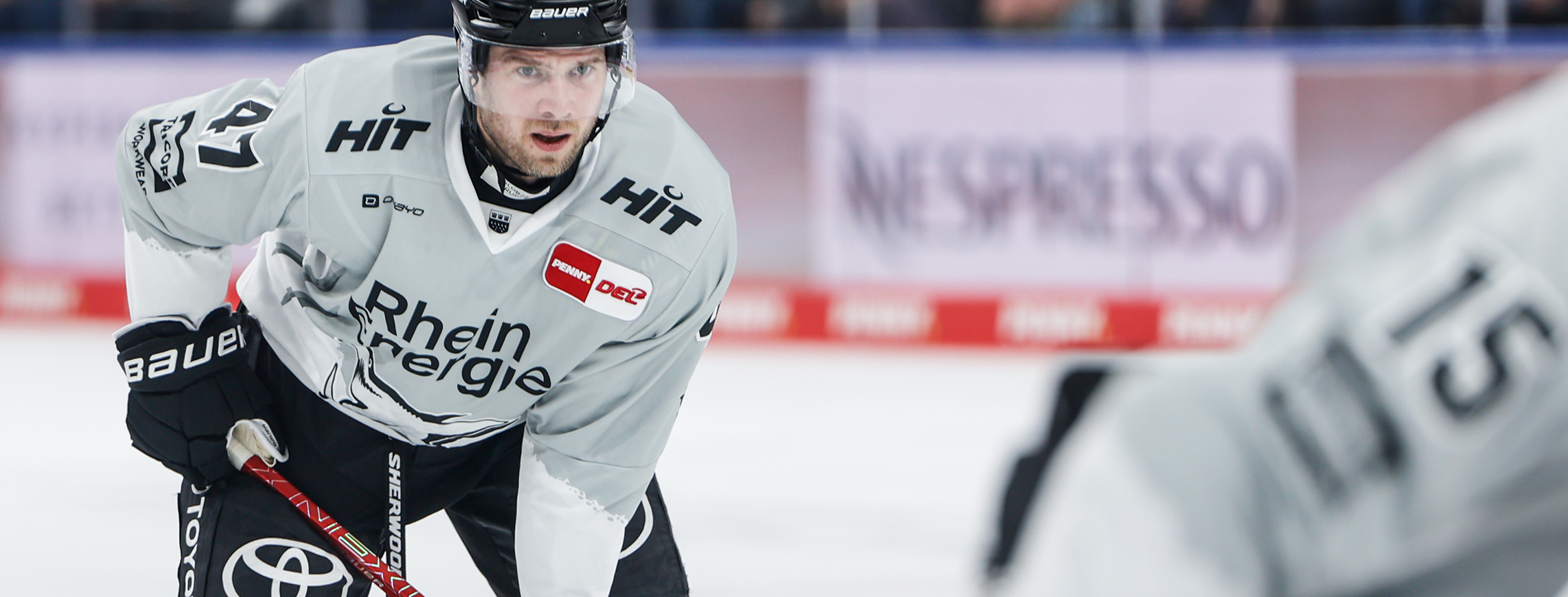 An ice hockey player in a gray Owayo jersey is focused during the game, with a teammate in the foreground and the boards in the background.