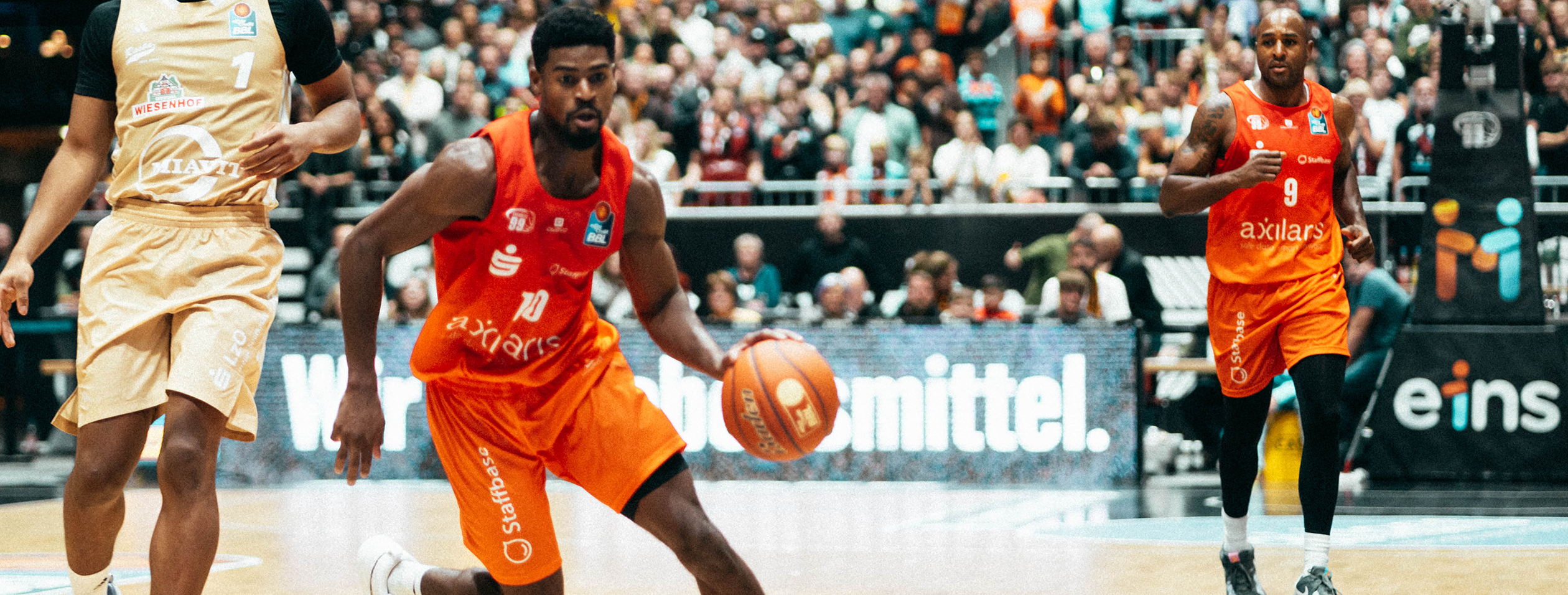 Two basketball players in orange Owayo jerseys during a game. One is dribbling the basketball, while the other runs alongside.