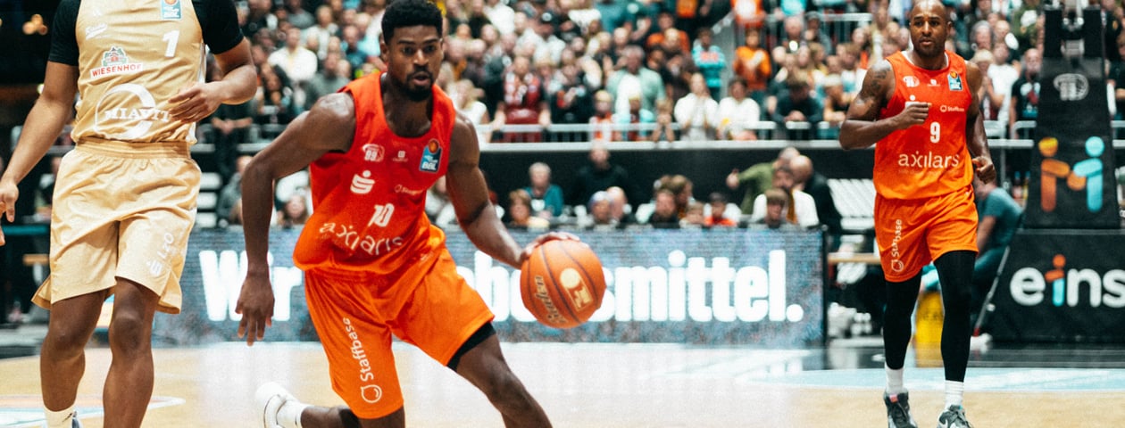 Two basketball players in orange Owayo jerseys during a game. One is dribbling the basketball, while the other runs alongside.