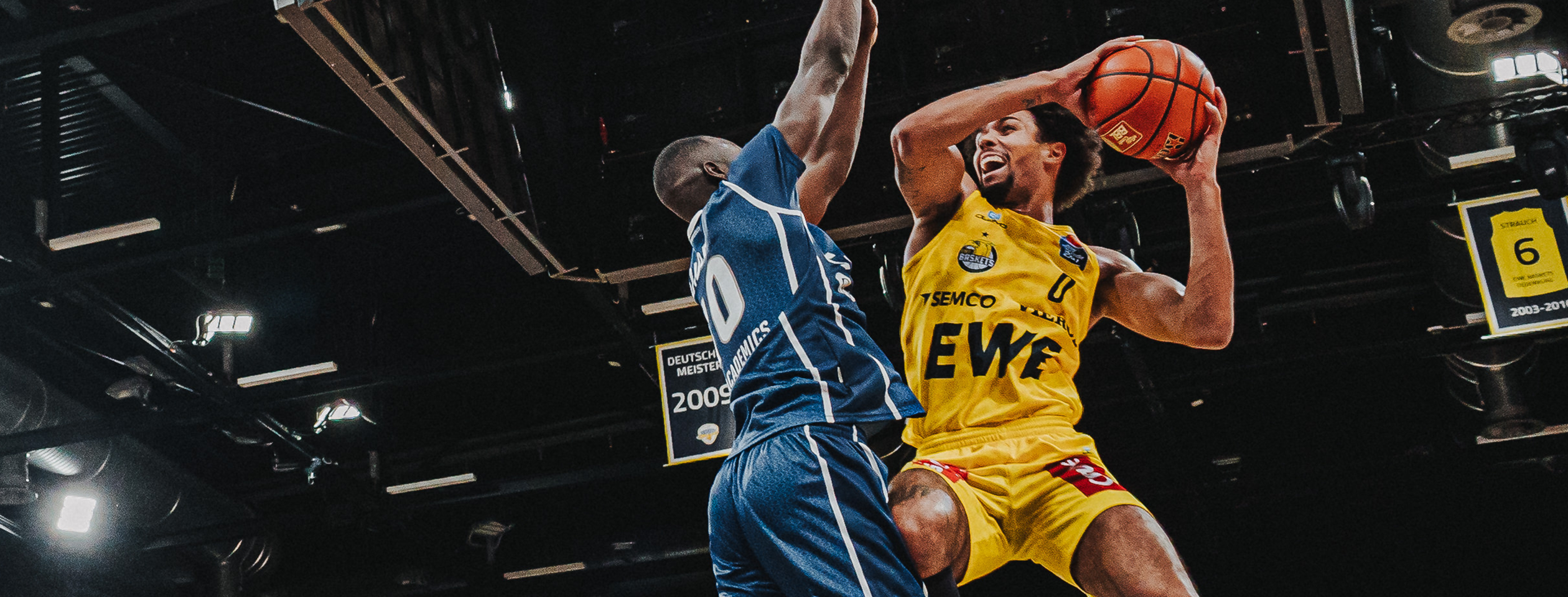 A basketball player in a yellow Owayo jersey taking a jump shot while an opponent in a blue jersey tries to block the shot