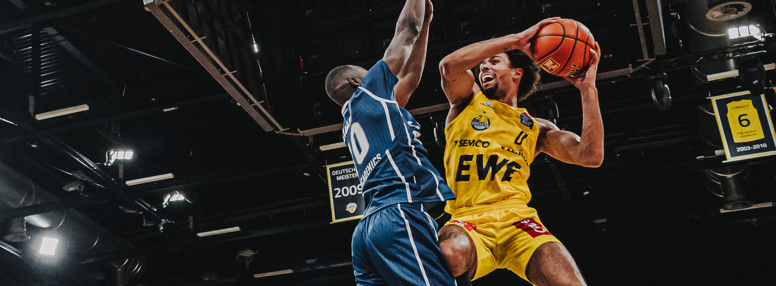 A basketball player in a yellow Owayo jersey taking a jump shot while an opponent in a blue jersey tries to block the shot