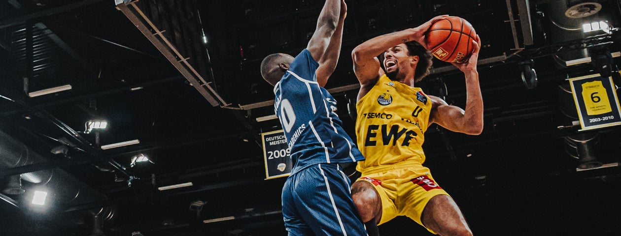 A basketball player in a yellow Owayo jersey taking a jump shot while an opponent in a blue jersey tries to block the shot