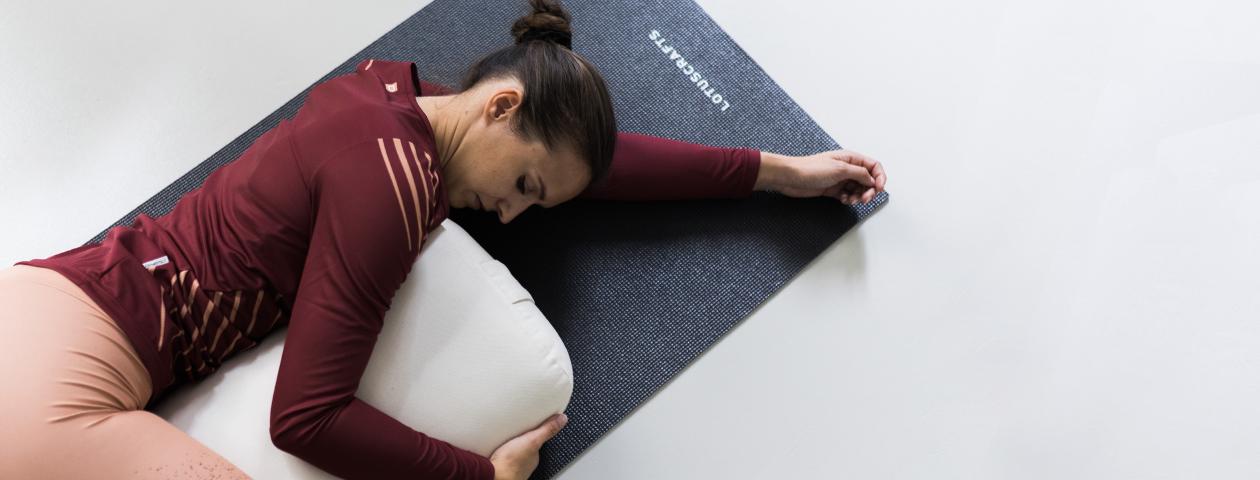 Woman stretching with yoga cushion on yoga mat wearing custom designed dark red yoga top and yoga pants in rose
